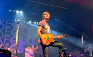 A male guitarist plays an orange guitar on stage, with band name 
