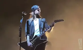 Guitarist with long blonde hair, beard, and baseball cap plays a black electric guitar on stage.
