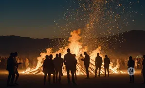 Silhouette of people gathered around a large bonfire at dusk with embers flying.