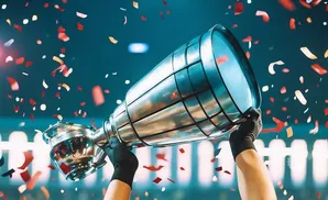 A person in black gloves holds up a trophy as red and gold confetti falls around them.