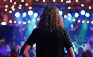 Person with long curly hair facing away from camera on a stage with blurred lights and audience.