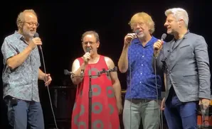 Four men on stage singing into microphones, two in casual shirts and two in more formal attire.
