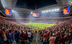 A football stadium filled with cheering fans at night, with a game in progress on the field and large screens displaying abstract art.