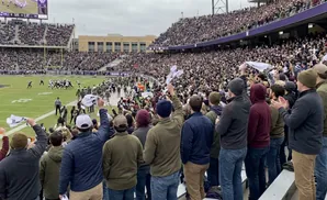 Fans in a football stadium wave white towels as players face off on the field under a cloudy sky.