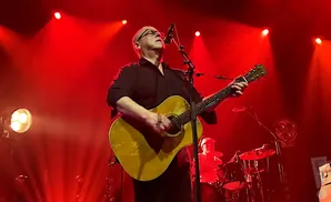 A bald man in glasses plays an acoustic guitar on a brightly lit red stage.