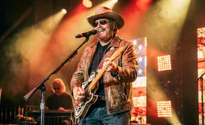 A man in a cowboy hat and leather jacket plays guitar and sings into a microphone on stage.