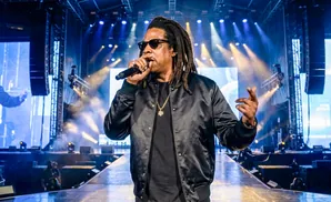 Jay-Z performs on stage wearing sunglasses, a black bomber jacket, and a gold chain.