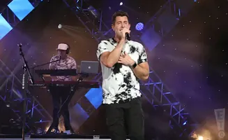 Singer on stage with a keyboard player, both wearing tie-dye shirts, with dramatic lighting.