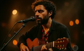A man with curly hair plays an acoustic guitar and sings into a microphone on a dimly lit stage.