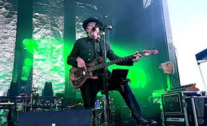A man in a hat and suit plays a bass guitar on a stage with green lighting.