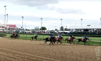Horse race with multiple horses and jockeys on a dirt track, with grandstands and lights in the background.