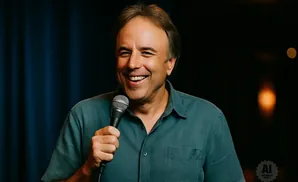 A smiling man with a microphone, wearing a teal button-down shirt, stands in front of a dark blue curtain.