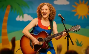 A woman with curly hair plays an acoustic guitar on stage, smiling into a microphone.