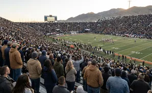 A football game viewed from the stands at sunset, with fans filling the stadium.