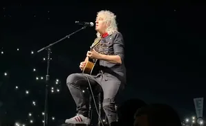A man with curly blonde hair, playing an acoustic guitar and singing into a microphone while sitting on a stool on a dark stage with many small bright l…