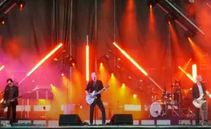 A band performs on a stage lit by bright orange lights, with guitars and drums visible.