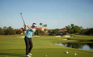 Golfer swings club on a lush green course with a large house in the background.
