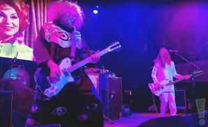 A band performs on stage under purple and blue lights. A guitarist in a colorful, eye-patterned outfit plays a white guitar.