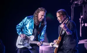 Two men playing guitars on stage. The man on the left wears a shiny blue jacket, and the man on the right wears a dark suit jacket.