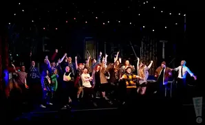 A diverse group of people on stage with their arms raised in celebration, with a dark background and small lights.