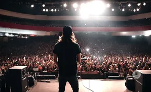 Musician on stage with back to audience in a packed, dimly lit arena with bright spotlights overhead.