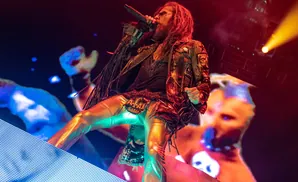 Rob Zombie performs on stage in a leather jacket with patches, metallic pants, and face paint.