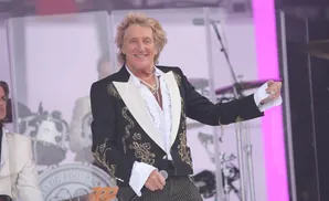 Rod Stewart performs on stage in a black sequined jacket and striped pants, wearing multiple necklaces.
