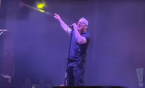 Singer with mohawk and glasses on stage, pointing and holding a microphone, under purple stage lights.
