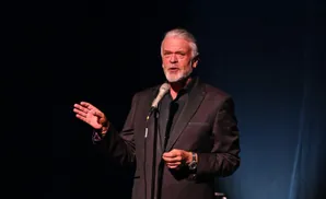 Man in a tuxedo speaks into a microphone, gesturing with his left hand.