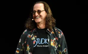 Geddy Lee of Rush smiles, wearing a floral shirt over a t-shirt that says 