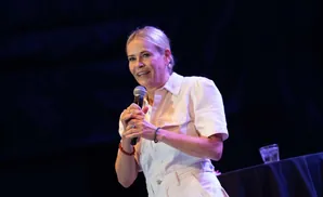 A woman with blonde hair smiles as she holds a microphone on a dark stage.