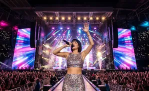 A woman with dark hair and red lipstick sings into a microphone, bathed in blue and pink stage lights.