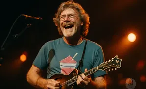 A man with glasses and a mustache plays a mandolin on stage, laughing and wearing a blue t-shirt with a slushy graphic.