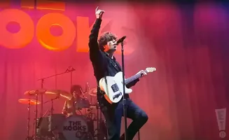 Singer of The Kooks performs on stage, raising one hand while playing a white electric guitar.