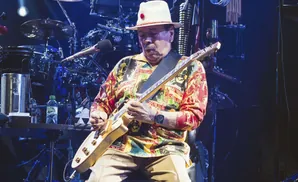 Carlos Santana plays a white guitar on stage, wearing a colorful shirt and white hat.