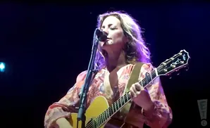 A woman with blonde, wavy hair sings into a microphone while playing an acoustic guitar.