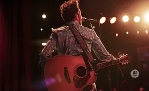 A musician plays an acoustic guitar on stage, illuminated by warm stage lights.