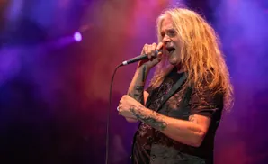 A male singer with long blonde hair, tattoos, and a sparkly vest sings into a microphone on a dimly lit stage.