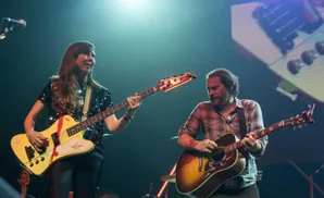 Two musicians play guitars on stage, bathed in blue and white stage lights.