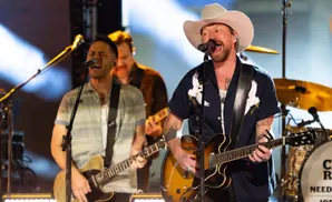 A country singer in a cowboy hat sings and plays guitar on stage with his band.
