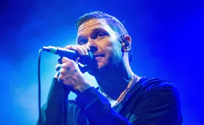 A man sings into a microphone on a blue-lit stage, wearing a chain necklace.