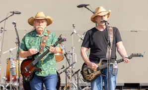 Two men in cowboy hats play guitars on stage, with drums in the background.