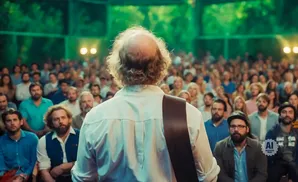 Bald man in white shirt plays guitar for an outdoor audience.