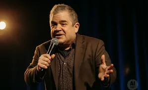Comedian Patton Oswalt holds a microphone and gestures with his hand while speaking on stage.