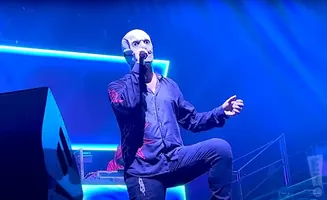 A performer in a white, mask-like helmet sings into a microphone on a stage bathed in blue light.