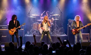 A rock band performs on stage with bright lights and fog. The band has a guitarist, bassist, singer, and drummer.