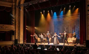 A cast performs on stage in a theater with an orchestra in the foreground and audience members in the lower seating.