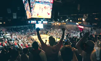 A crowd of cheering basketball fans with arms raised in excitement watch a game on a large screen in a stadium.