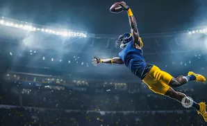 An American football player in mid-air, catching a pass with a stadium lit up behind him.