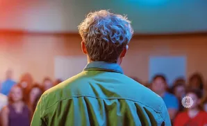 Man with curly hair addresses an audience from behind, bathed in dramatic red and blue stage lighting.